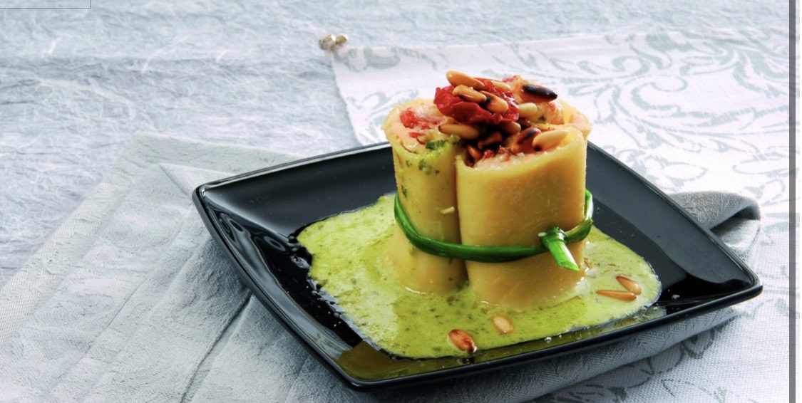 A cylindrical pasta dish sits on a black plate with green sauce and chopped nuts, photographed in bright natural light.