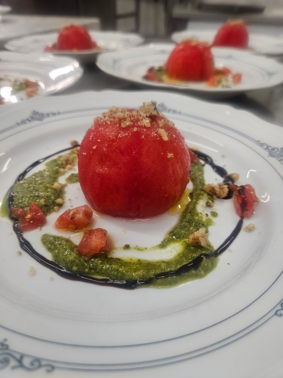 A red spherical plated dessert surrounded by green sauce, dark sauce and nuts, with other plates in the background.
