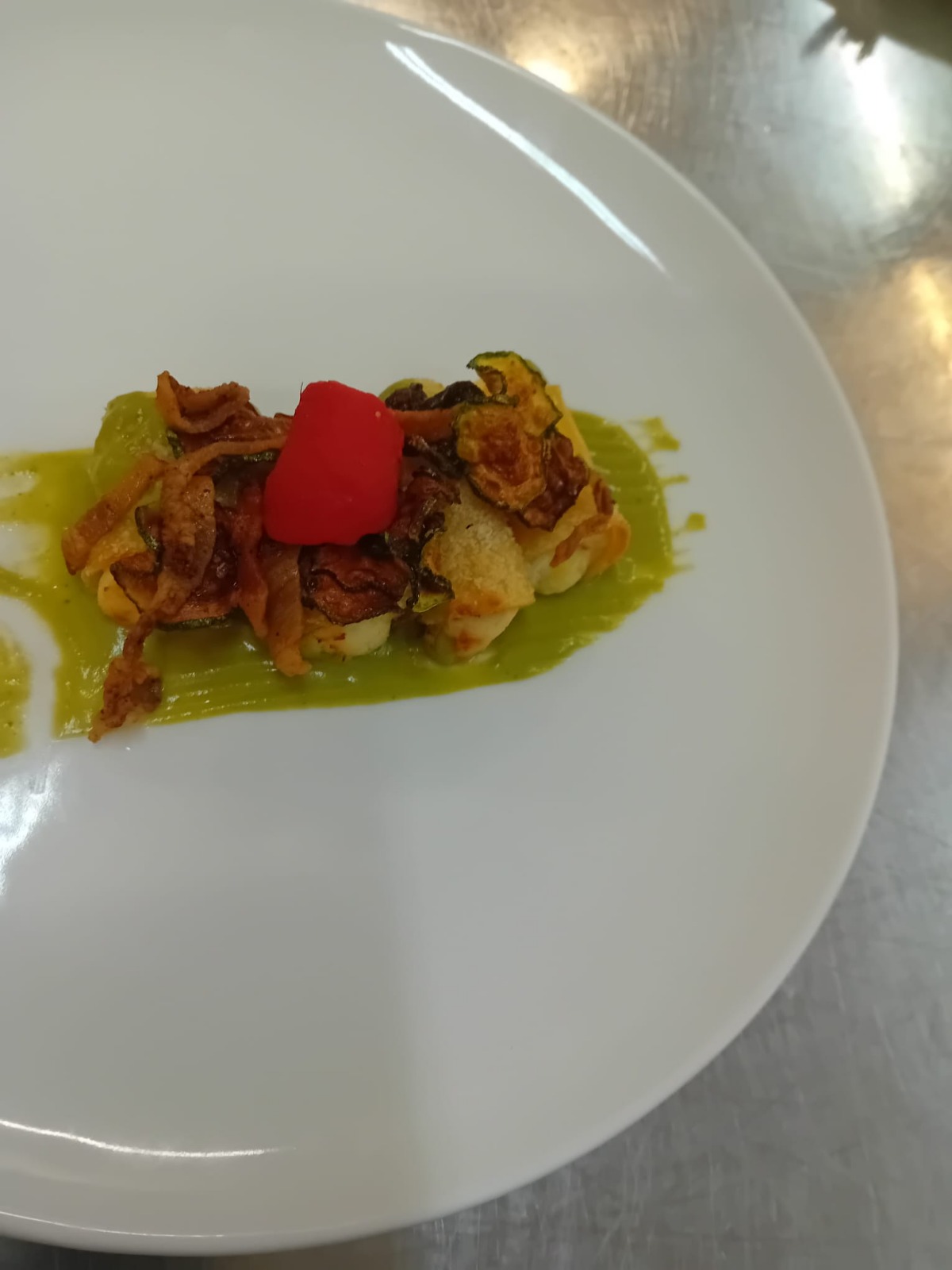 A plated entrée with vegetables and crisp toppings on green sauce, photographed from above under indoor lighting.
