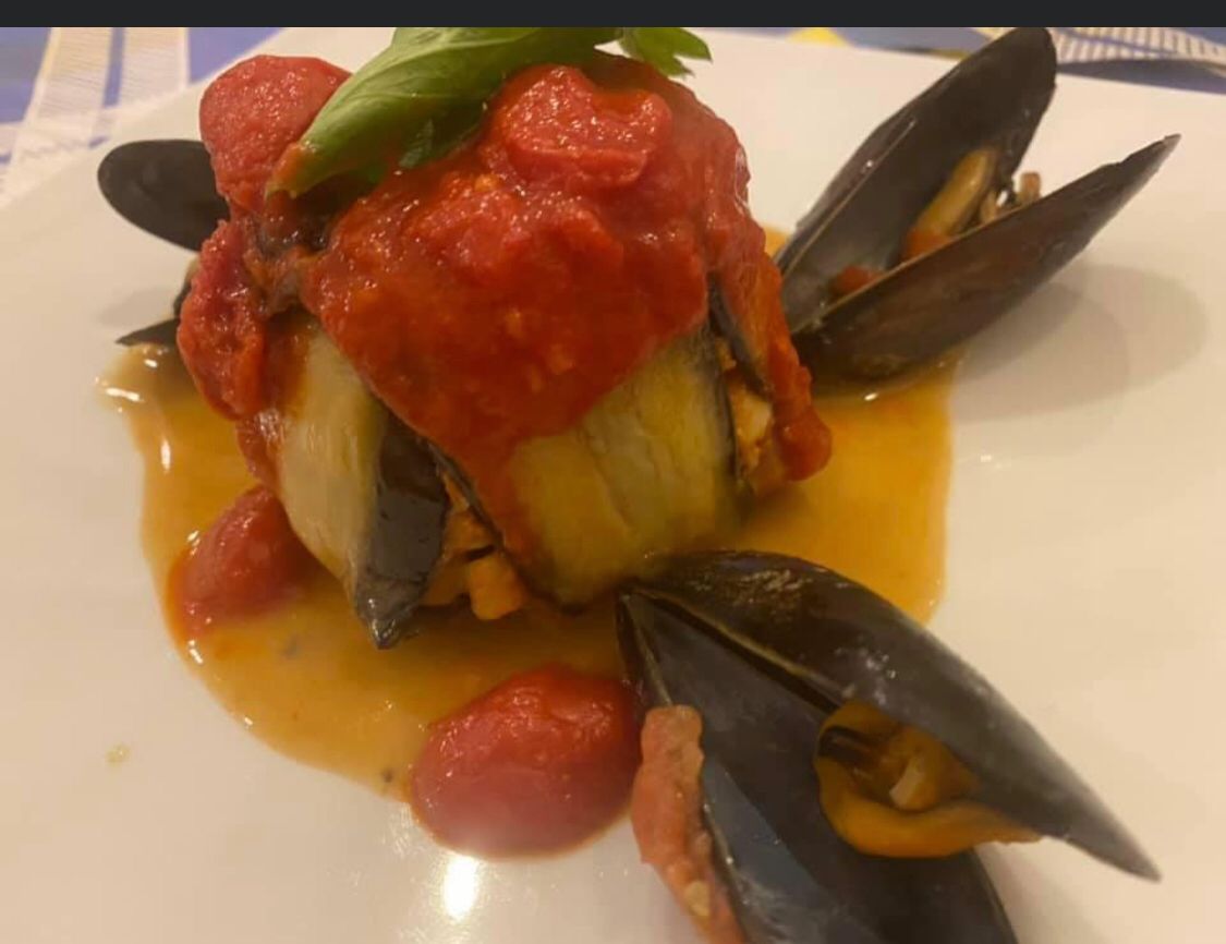 Close-up of a plated seafood dish with mussels, tomato sauce and vegetables on a white plate, warmly lit.