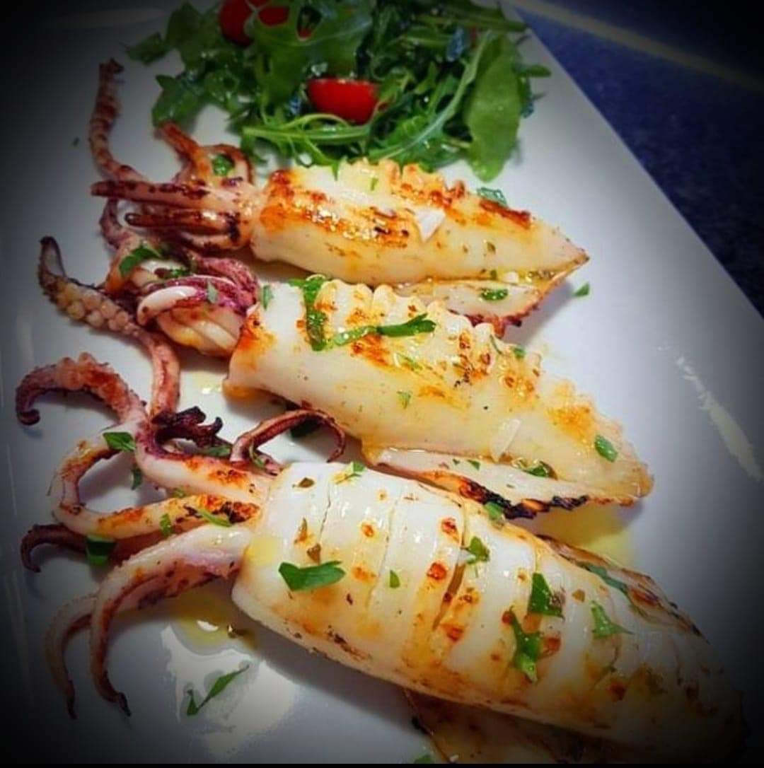 Grilled squid arranged on a white plate with greens and herbs, softly lit.