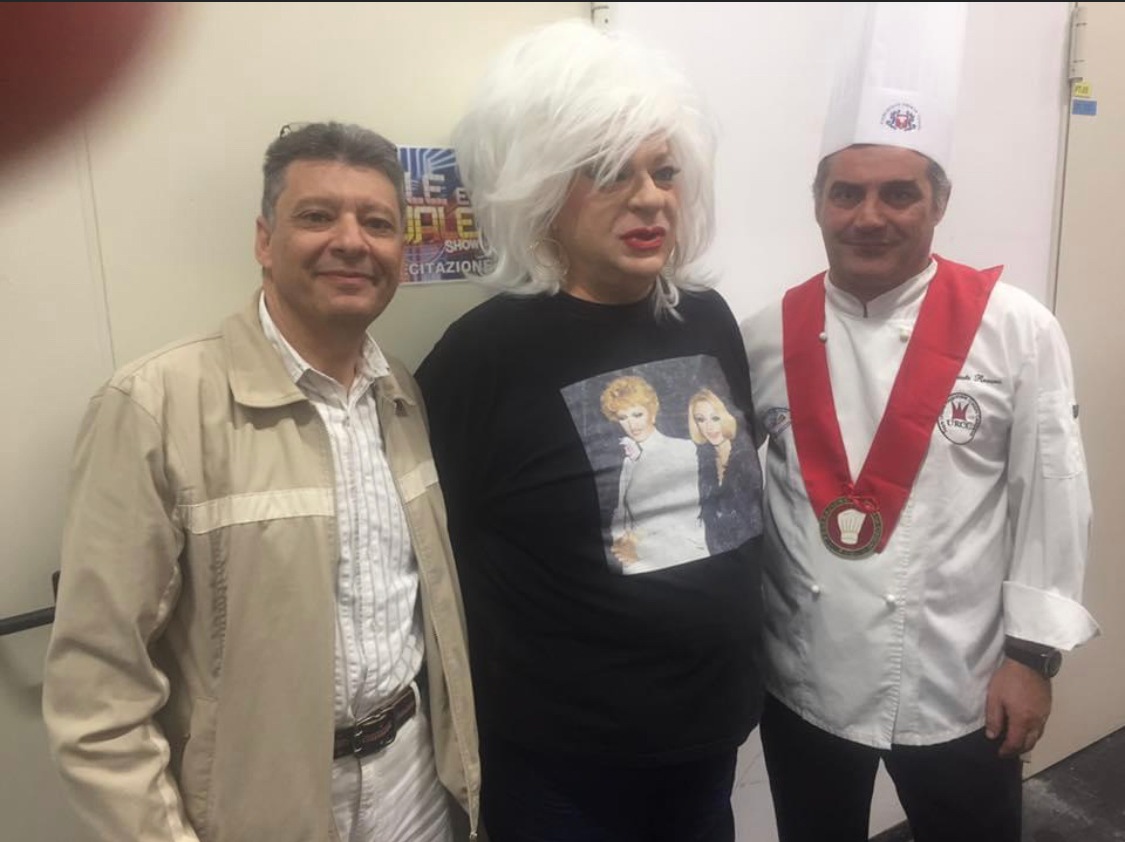 Three people pose indoors, including one person in a chef hat and ceremonial sash, under bright overhead lighting.